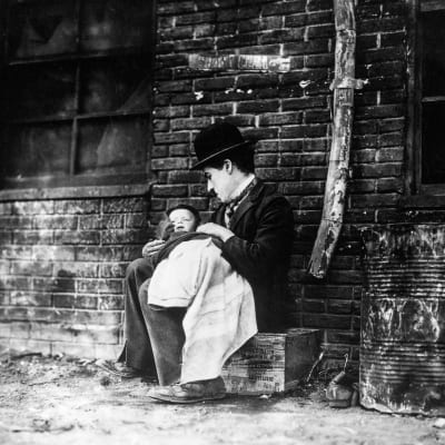 Knallipäinen kulkuri (Charlie Chaplin) istuu pakkilaatikolla rähjäisellä seinänvierustalla pieni lapsi sylissään. Kuva elokuvasta Chaplinin poika.