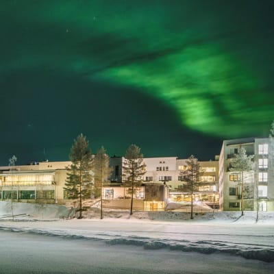 Talvinen maisema, jossa puiden takana kylpylärakennus. Taivaalla revontulia.