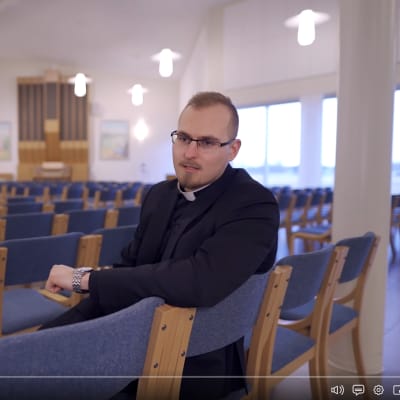 En präst sitter på en stol i en stor sal och pratar.