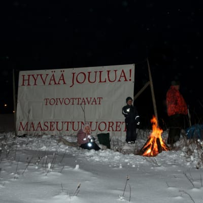 Nuotio palaa talvisessa iltamaisemassa. Taustalla on iso kyltti, joka toivottaa hyvää joulua.
