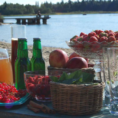 Ett bord uppdukat med en bricka med röda vinbär, en skål hallon, en skål jordgubbar, en korg äppel, en korg svarta vinbärsblad, en karaff med appelsinjuice, några flaskor cider, en karaff med svagdricka och några highball-glas, på en sandstrand med havet