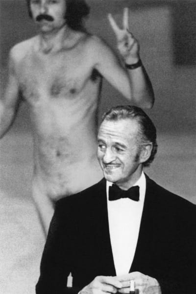 Svartvit bild där Robert Niven står i förgrunden iklädd smoking och ser glad ut, bakom honom skymtar man en naken Robert Opel som visar segertecknet.