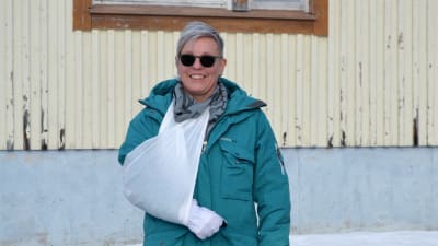 Porträttfoto av kvinna med grönblå jacka, svarta solglasögon, ena armen i mitella. Hon tittar in i kameran och ler stort. Bakom en träbyggnad som är beigemålad.