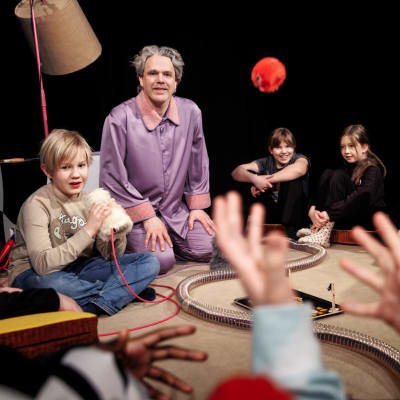 En grupp barn sitter på ett golv omkring en man i en lavendelfärgad pyjamas. 