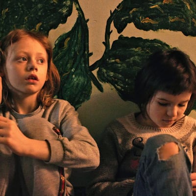 Kaksi nuorta tyttöä istuu seinää vasten, johon on maalattu kasveja. Harmaan collegeen pukeutunut punahiuksinen tyttö katsoo oikealle, neulepaitaan pukeutunut tyttö katsoo alas.