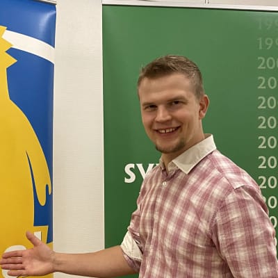 Kielilähettiläs Jaan Siivonen ojentaa kättä suuressa julisteessa olevalle Svenska veckanin tunnushahmolle, Svenni-lohikäärmeelle, joka toivottaa päivää - gudaa.