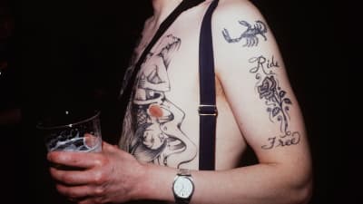 En man med vaken överkropp som är tatuerad. Han håller i en öl. Huvudet syns inte, bilden är avskuren vid axlarna.