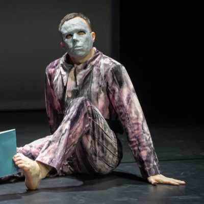 Koreografen Carl Knif sitter på golvet med en mask på ansiktet och ett blått häfte mallen tårna.