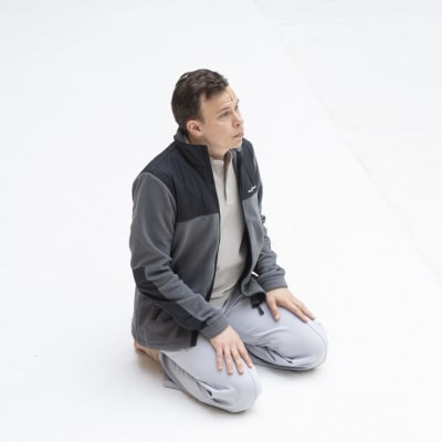 Ruskeatukkainen mies (tanssija Carl Knif) harmaissa verkkareissa ja fleecepuserossa istuu polvillaan lattialla vasten täysin valkoista taustaa ja katsoo yläviistoon oikealle.