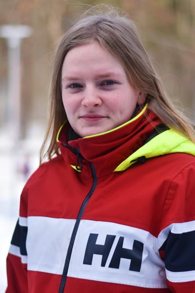 Ung kvinna med långs ljust hår och sportjacka i rött, vitt och blått ser in i kameran och småler. I bakgrunden träd, lyktstolpar och snö på marken.