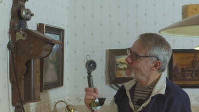 En äldre man med glasögon står i ett rum, håller i luren till en gammal telefon hägnd på väggen. 