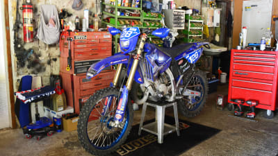 En motorcykel står inne i ett garage. På väggen bakom cykeln syns olika verktyg och annat som behövs för att reparera den.