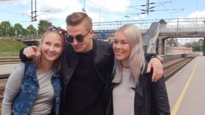 Tre unga vuxna poserar avslappnat på en järnvägsperrong. Det är två kvinnor och en man. Man ser räls och i bakgrunden en bro för biltrafik. Det är sommar..