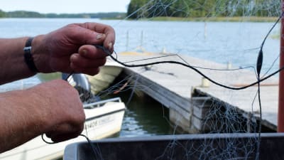 En par gamla händer håller i ett fiskenät, somrigt hav och båtar i bakgrunden.