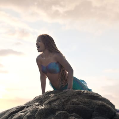 Sjöjungfrun Ariel i Halle Baileys gestlat sjunger på en klippa med solnedgång i bakgrunden.