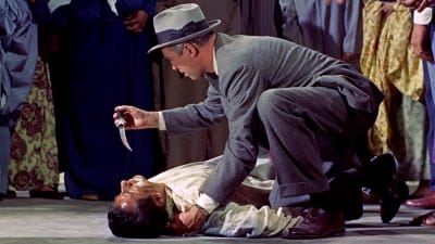Länsimaiseen pukuun pukeutunut hattupäinen mies (näyttelijä James Stewart) katsoo kädessään olevaa veristä tikaria maassa makaavan murhatun miehen ruumiin ylle kyyristyneenä; taustalla näkyy pohjoisafrikkalaisittain pukeutuneita ihmisiä. Kuva elokuvasta Mies joka tiesi liikaa.