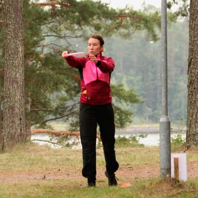 Katri Koponen seisoo frisbee ojennetuissa käsissään frisbeegolfradalla ja valmistautuu heittämään, taustalla mäntymetsää ja lampi.