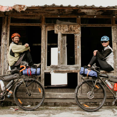 Ilkka ja Tuija Kauppinen istuvat ulkomaalaisen talonrähjän edustalla retkipyöräilyvarusteissa.