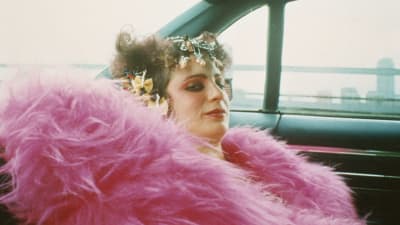 Nan Goldin sitter i baksätet i en bil och ser rakt in i kameran, hon har en enorm rosa lurvig och fluffig rock och smycken i håret. 