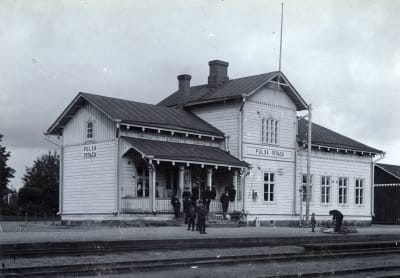 Mustavalkoinen arkistokuva vanhasta puisesta rautatieasemasta, edessä henkilöitä odottamassa, 1900-luvun alkua.