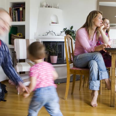 Äiti ja lapsia kotona kahvipöydässä, pikkulapset juoksevat lattialla.