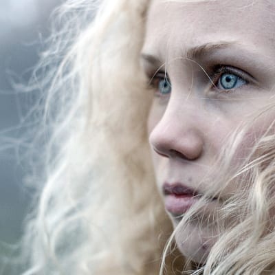 Karaktären Tyra i närbild med lockigt hår och kallblå ögon.