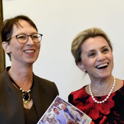 En leende Sari Essayah och en leende Päivi Räsänen, som håller i en broschyr, framför Kristdemokraternas logga.