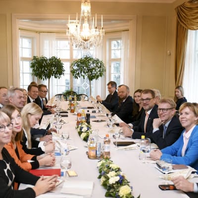 Representanter för alla partier sitter samlade i statsministerns tjänstebostad kring ett bord med vit duk och takkrona.
