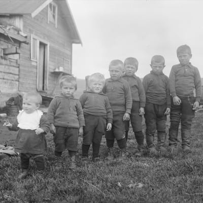 Seitsemän lasta seisoskelevat mustavalkoisessa kuvassa rivissä. 