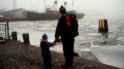 En man och en pojke står i hamnen i Helsingfors och ser ut över vattnet på en ångbåt
