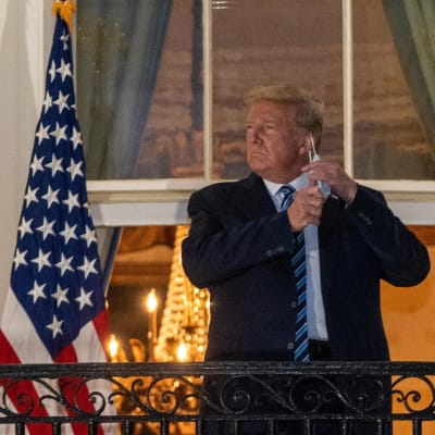 Donald Trump står på Vita husets trappa framför amerikanska flaggor och tar av sig sitt munskydd när han kommer hem från sjukhuset.
