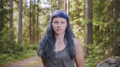 Hantverkskonstnären Elena Bondar i närbild. I bakgrunden en tät granskog. Elena Bondar har långt, blåfärgat hår med pannlugg.