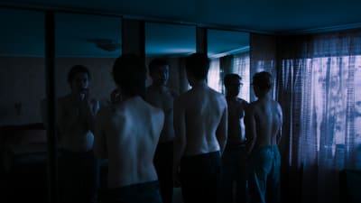 Tre pojkar med bar överkropp står framför en stor spegel i ett halvmörkt rum och tittar på sig själva.