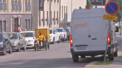 Ett cykelbud med gul skjorta sitter i sin cykel med pedalerna framför sig och en stor gul låda bakom sig. Han cyklar förbi en av Postens bilar.