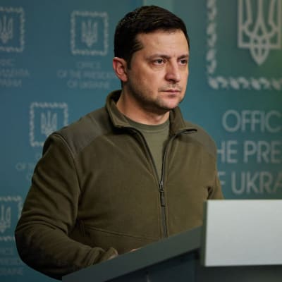 Volodymyr Zelenskyj fotograferad från sidan när han står vid ett talarpodium och pratar. I bakgrunden Ukrainas flagga.