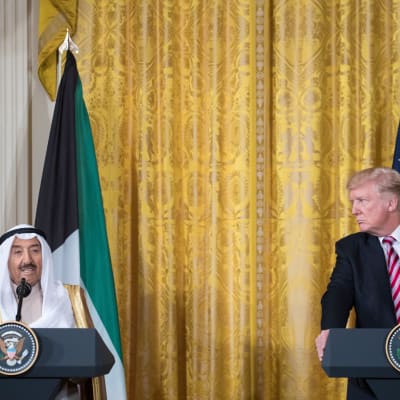 Donald Trump katsoo kohti miestä, joka puhuu mikrofoniin. Taustalla yhdysvaltain ja Kuwaitin liput. 