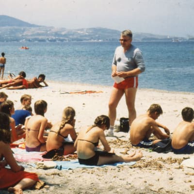Opettaja Juhani Vahtokari seisoo shortsiasuisena oppilaisen edessä hiekkarannalla jossakin Välimeren alueella.