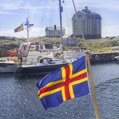 En båt på vatten. Från båten vajar Ålands flagga. I bakgrunden fler båtar och ett hus.