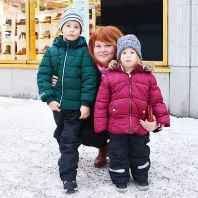 En kvinna sitter på knä och håller armarna om två barn, en flicka och en pojke. Familjen är utomhus i en snöig stadsmiljö.