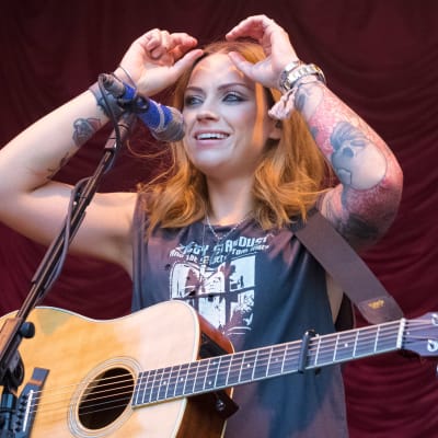 Amy Macdonald har en akustisk gitarr kring halsen och står bakom en mikrofonställning.