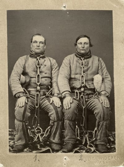 Två fångar i vadmalsuniformer och med tunga fängsel om hals, midja och ben.