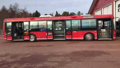 En röd buss som står parkerad på rött grus utanför en röd hall.