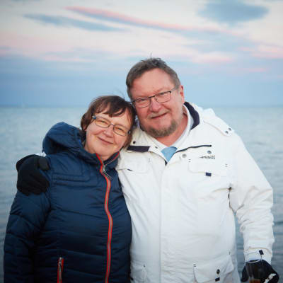 Raili ja Juha M. Venäläinen poseeraavat hymyillen ulkoiluvaatteissa rannalla taustallaan avoin ulappa.