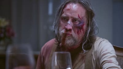 En blodig och blåslagen Rob (Nicholas Cage) sitter vid ett bord och ser allvarlig ut.