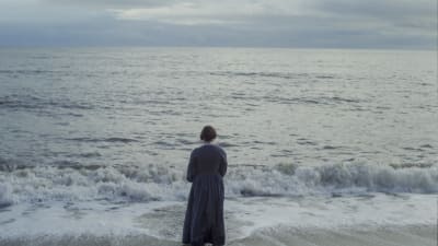 Mary Anning (Kate Winslet) står vid vattenbrynet vid havet och ser mot horisonten. 