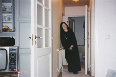 Iiu Susiraja seisoo kotona eteiskäytävässä mustiin vaatteisiin pukeutuneen ja nojaa olkapäällään seinään.