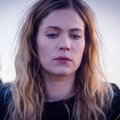 Sofia Karppi (Pihla Viitala) katsoo vakavana jotain alaspäin. Hänen hiuksensa ovat kiharaiset ja auki. Takana kollega Sakari Nurmi (Lauri Tilkanen)