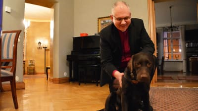 En medelålders man i svart kavaj står på knä och klappar en brun labrador. Hunden tittar in i kameran.