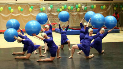 En grupp gymnaster med bollar i händerna knäböjer på scen.