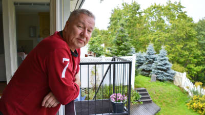 En man i röd tröja lutar sig över ett balkongräcke och tittar in i kameran.
